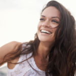 Brunette woman in a white tanktop smiles because she has good oral health and overall health