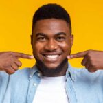Man with no cavities points to his healthy smile while standing against a yellow background