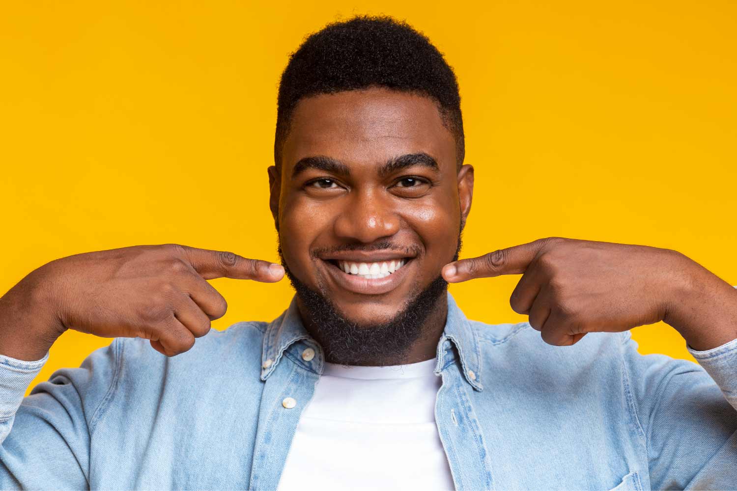 Man with no cavities points to his healthy smile while standing against a yellow background