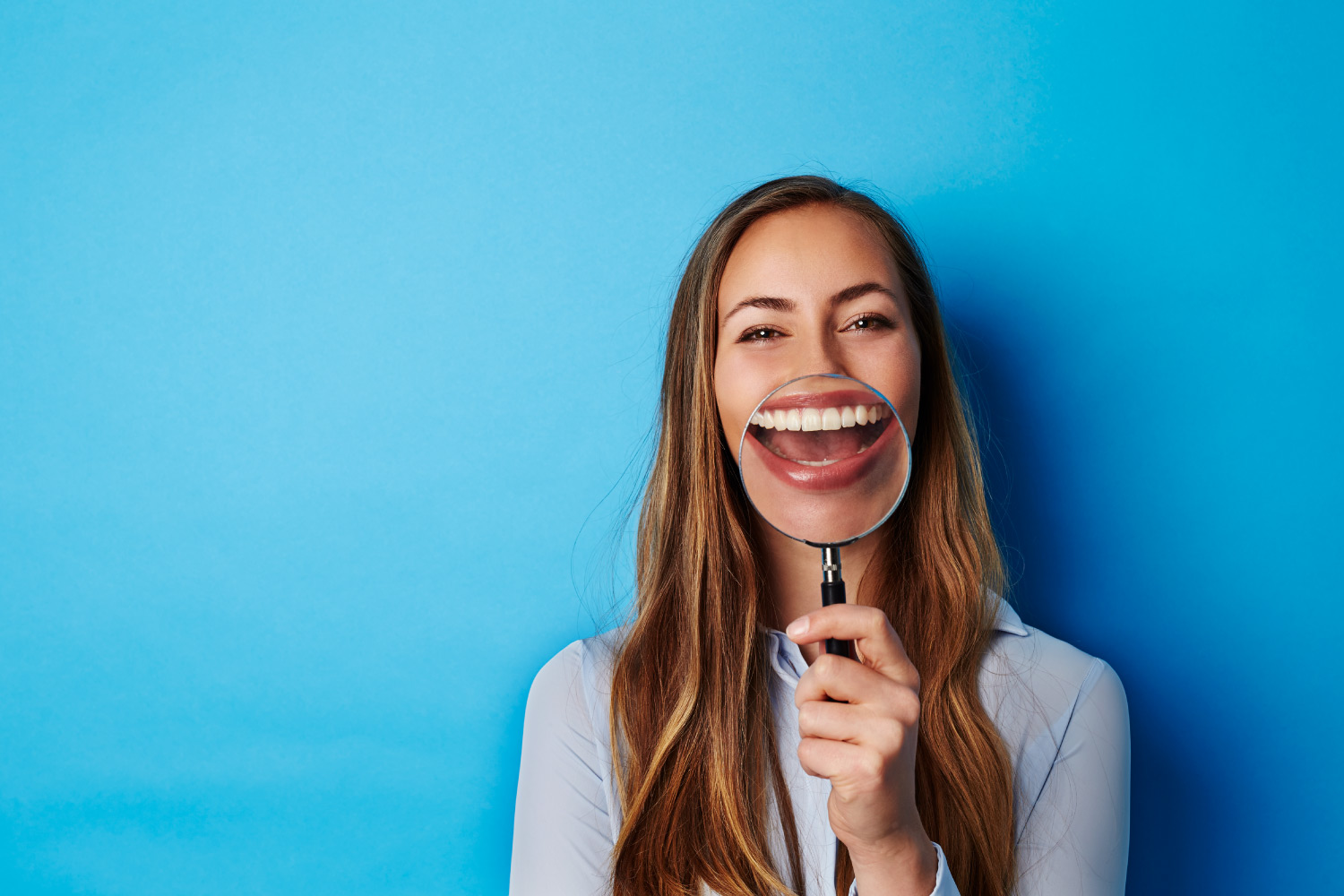 Brunette woman holds a magnifying glass up to her teeth and gums against blue background