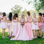 Photo of the back of a line with a bride surrounded by bridesmaids dressed in pink out in a field.