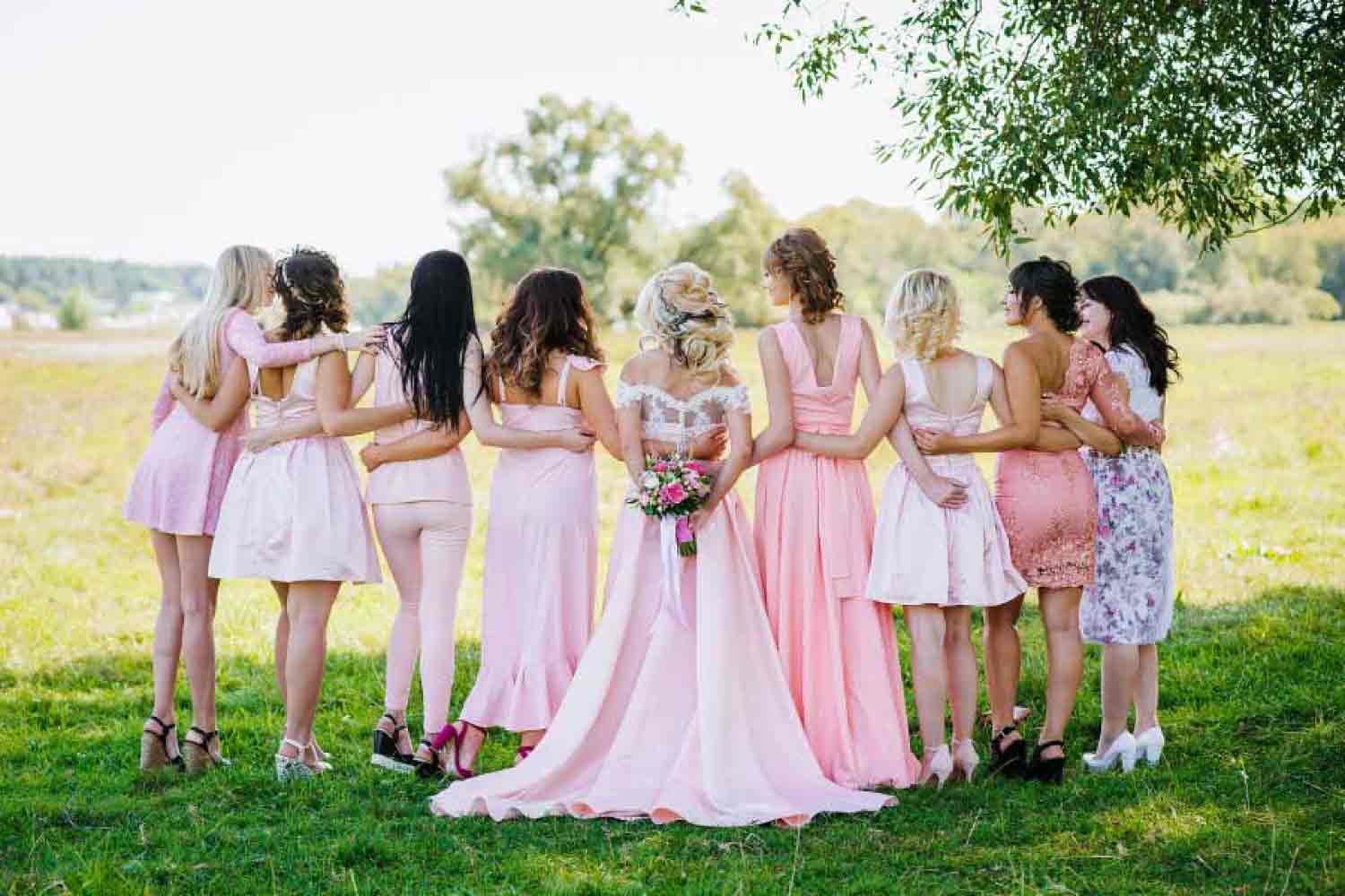 Photo of the back of a line with a bride surrounded by bridesmaids dressed in pink out in a field.
