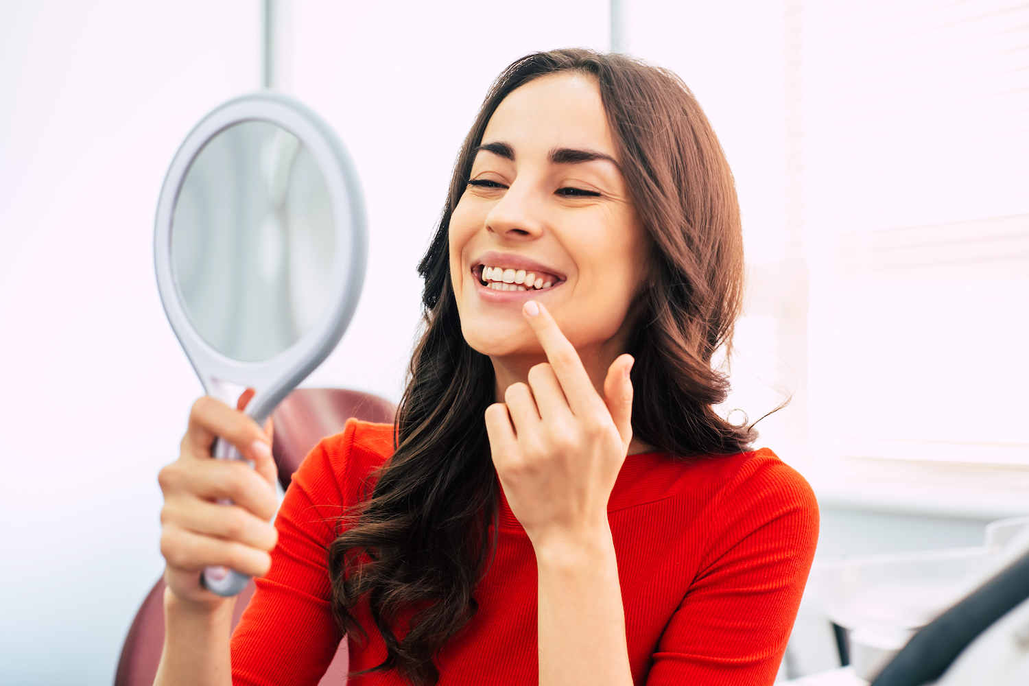 Smiling woman with long curly hair in the dental chair is using a mirror to see the result of dental work